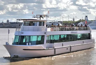 O navio de cruzeiro Burdigala II navegando no rio Garona em Bordéus, com passageiros no convés superior e o horizonte histórico da cidade ao fundo.