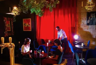 Interior do Théâtre des Chartrons em Bordéus com três pessoas num elegante bar com paredes vermelhas, mobiliário azul, iluminação sofisticada e decoração vegetal.