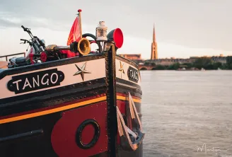 O barco Tango amarrado no rio Garona em Bordéus, decorado com cores vibrantes, uma bandeira vermelha, instrumentos musicais e enfeites, com a Basílica de Saint-Michel visível ao fundo.