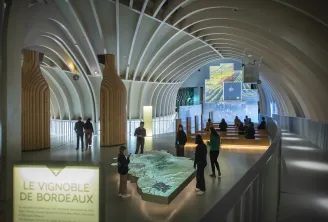 Interior da Cité du Vin em Bordéus com arquitetura moderna de teto abobadado branco brilhante, visitantes observando uma maquete iluminada da vinha de Bordéus e ecrãs de exposição nas paredes.