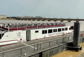 O Maddalena, um iate de cruzeiro fluvial branco e vermelho, amarrado a um cais em Bordéus com o rio Garona e a Ponte de Pierre visíveis ao fundo.