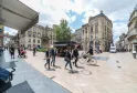 Praça movimentada em Bordéus com peões, ciclistas e lojas, ladeada por edifícios estilo haussmanniano sob céu nublado.