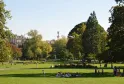 Vista do Jardim Público de Bordéus com relvados verdes, árvores majestosas, visitantes a relaxar, um monumento com estátua ao fundo e edifícios históricos sob céu limpo.